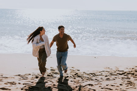 A smiling couple enjoys a carefree run on a sandy shore, waves gently breaking in the backgroundの写真素材
