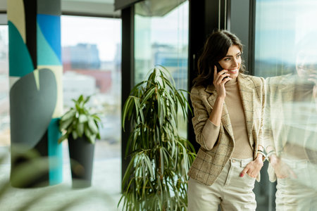 Contemplative woman using mobile phone and gazes out of a window, with a thoughtful expression and city views behind herの写真素材