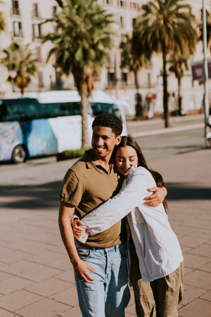 Young couple holds hands and smiles as they walk down a sunlit path in Barcelona, framed by palmsの写真素材