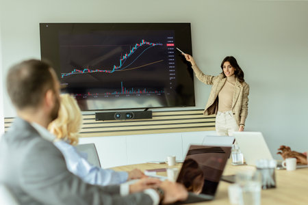Businesswoman points at growth trends on a screen during a financial meeting, with attendees listening intentlyの写真素材