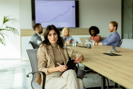 Focused businesswoman with a notepad ponders during a team meeting, with colleagues engaged in discussion in the backgroundの写真素材