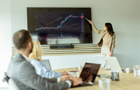 Businesswoman points at growth trends on a screen during a financial meeting, with attendees listening intentlyの写真素材