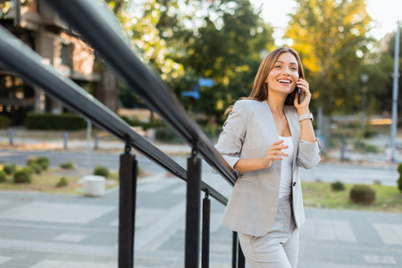 Woman strolls outdoors in a chic gray suit, animatedly chatting on her phone. The natural light enhances her radiant smile, surrounded by beautiful greeneryの写真素材