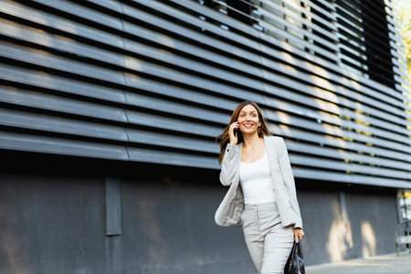 Walking with purpose, a business woman in a stylish outfit enjoys a phone conversation on a bright day, showcasing her confident demeanor against a sleek architectural backdropの写真素材