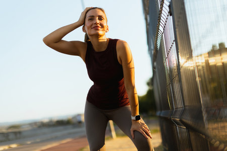 Woman pauses during her evening workout, wiping her brow as the sun sets, surrounded by a tranquil jogging path and a softly lit landscapeの写真素材