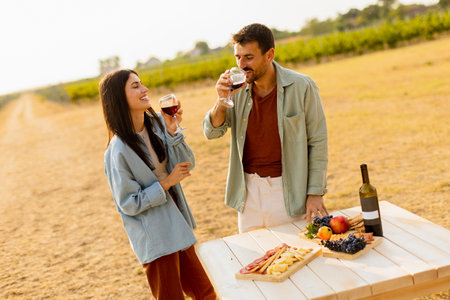 Couple relaxes at a rustic table in a sunlit vineyard, savoring wine and gourmet snacks. They share laughter and joy, surrounded by lush vines and the warmth of the eveningの写真素材