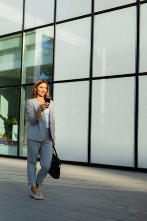 Buzsiness woman walks purposefully along a sleek glass facade, engaged with her phone. The sunlight highlights her professional look, embodying a blend of elegance and modernityの写真素材