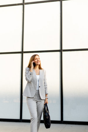 Professional woman strolls confidently along a sleek glass structure, her expression joyful as she chats on the phone, dressed in a stylish gray business suitの写真素材