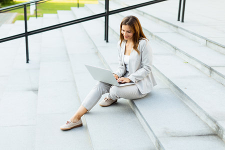 Seated on the steps, a woman in business attire focuses intently on her laptop, blending professionalism with the vibrant energy of the outdoors during a sunny dayの写真素材