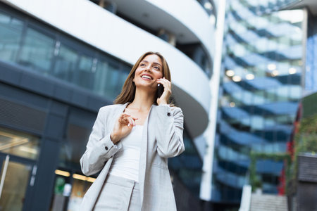 A professional woman engages in a cheerful phone conversation, exuding confidence as she strolls by modern buildings, basking in the warmth of a bright dayの写真素材