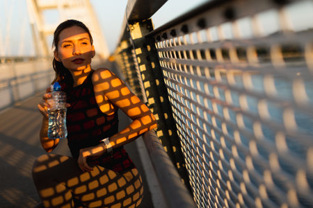 On a bright day, a woman rests on a bridge, holding a water bottle as sunlight filters through the railing, casting intricate shadows over herの写真素材