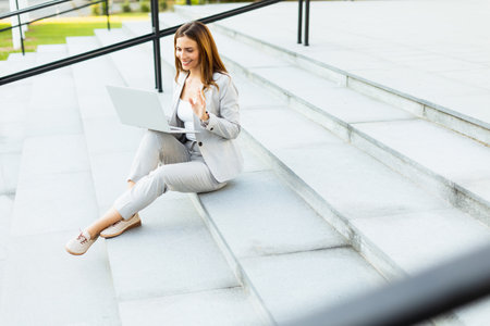 Seated on the steps, a woman in business attire focuses intently on her laptop, blending professionalism with the vibrant energy of the outdoors during a sunny dayの写真素材