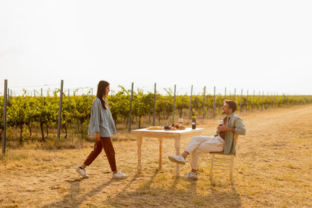 Couple relaxes at a rustic table in a sunlit vineyard, savoring wine and gourmet snacks. They share laughter and joy, surrounded by lush vines and the warmth of the eveningの写真素材