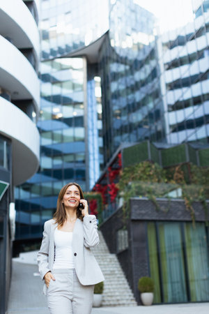 A professional woman engages in a cheerful phone conversation, exuding confidence as she strolls by modern buildings, basking in the warmth of a bright dayの写真素材