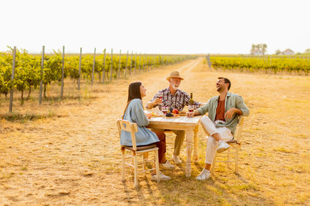 Three friends gather around a rustic table in a vineyard, enjoying wine, fresh fruits, and each other's company on a sunny day filled with joy and laughterの写真素材