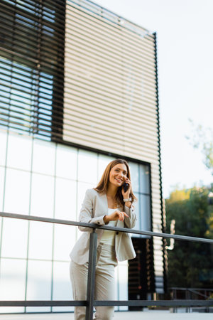 Cheerful business woman stands on a balcony, engaged with her smartphone against a backdrop of modern architecture, soaking in the warm sunlight and vibrant atmosphereの写真素材
