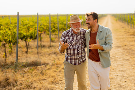 Walking along a dusty path in a sunny vineyard, two men engage in lighthearted conversation, their camaraderie evident against the backdrop of sprawling grapevinesの写真素材