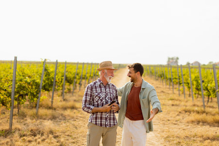 Walking along a dusty path in a sunny vineyard, two men engage in lighthearted conversation, their camaraderie evident against the backdrop of sprawling grapevinesの写真素材
