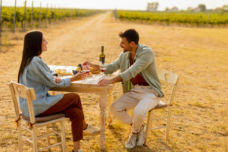 Couple relaxes at a rustic table in a sunlit vineyard, savoring wine and gourmet snacks. They share laughter and joy, surrounded by lush vines and the warmth of the eveningの写真素材