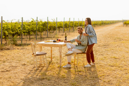 Couple relaxes at a rustic table in a sunlit vineyard, savoring wine and gourmet snacks. They share laughter and joy, surrounded by lush vines and the warmth of the eveningの写真素材