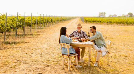 Three friends gather around a rustic table in a vineyard, enjoying wine, fresh fruits, and each other's company on a sunny day filled with joy and laughterの写真素材