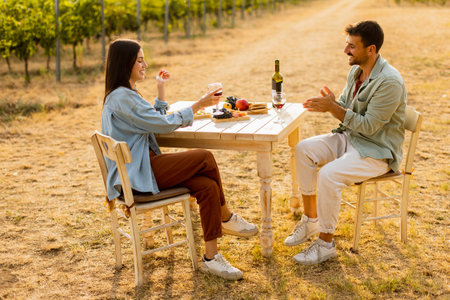Couple relaxes at a rustic table in a sunlit vineyard, savoring wine and gourmet snacks. They share laughter and joy, surrounded by lush vines and the warmth of the eveningの写真素材