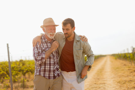 Walking along a dusty path in a sunny vineyard, two men engage in lighthearted conversation, their camaraderie evident against the backdrop of sprawling grapevinesの写真素材