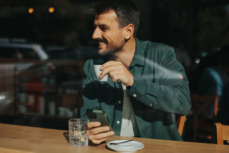 Sitting at a wooden table, a man enjoys his coffee while using mobile phone and gazing out a window, capturing the essence of solitude and reflection in a warm cafe atmosphereの写真素材