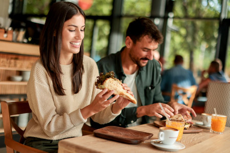 Couple shares laughter and delicious food while sitting at a wooden table in a bright cafe, surrounded by greenery and other patrons enjoying their mealsの写真素材