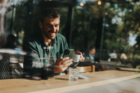 Sitting at a wooden table, a man enjoys his coffee while using mobile phone and gazing out a window, capturing the essence of solitude and reflection in a warm cafe atmosphereの写真素材