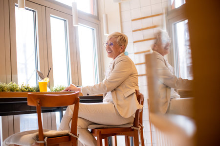 An older woman with stylish glasses smiles while working on her laptop at a cafÃ©, surrounded by greenery and sipping a refreshing drink, creating a lively atmosphereの写真素材