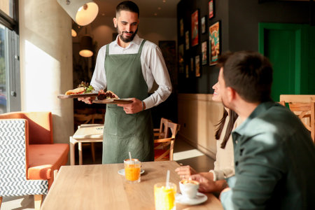 In a lively cafe filled with colorful artwork, a waiter presents fresh vegetarian meals to a smiling couple enjoying their afternoon together over refreshing drinksの写真素材