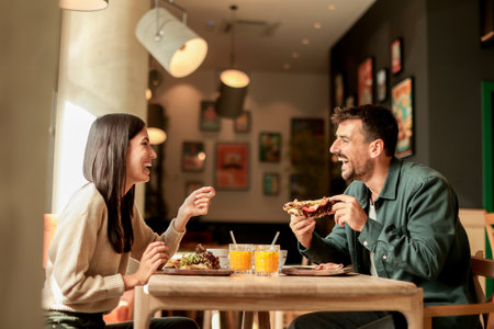 Couple shares laughter and delicious food while sitting at a wooden table in a bright cafe, surrounded by greenery and other patrons enjoying their mealsの写真素材