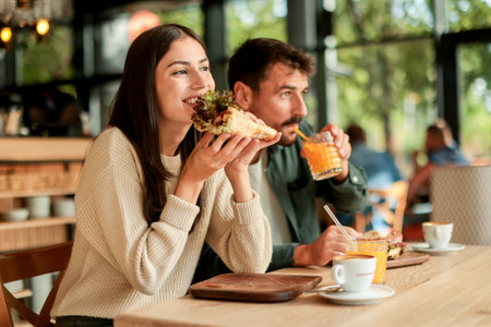 Couple shares laughter and delicious food while sitting at a wooden table in a bright cafe, surrounded by greenery and other patrons enjoying their mealsの写真素材
