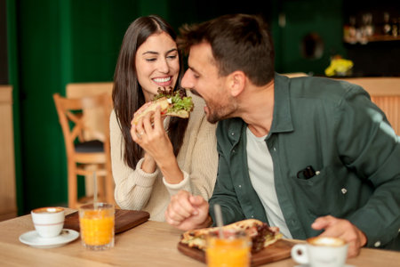 Couple shares laughter and delicious food while sitting at a wooden table in a bright cafe, surrounded by greenery and other patrons enjoying their mealsの写真素材