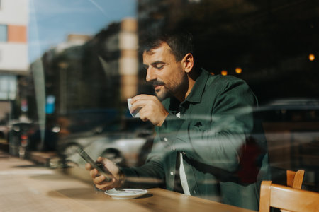 Sitting at a wooden table, a man enjoys his coffee while using mobile phone and gazing out a window, capturing the essence of solitude and reflection in a warm cafe atmosphereの写真素材