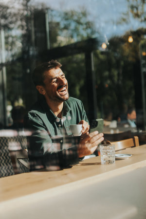 Sitting at a wooden table, a man enjoys his coffee while using mobile phone and gazing out a window, capturing the essence of solitude and reflection in a warm cafe atmosphereの写真素材
