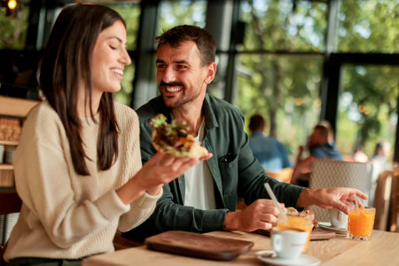 Couple shares laughter and delicious food while sitting at a wooden table in a bright cafe, surrounded by greenery and other patrons enjoying their mealsの写真素材