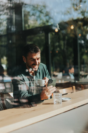 Sitting at a wooden table, a man enjoys his coffee while using mobile phone and gazing out a window, capturing the essence of solitude and reflection in a warm cafe atmosphereの写真素材