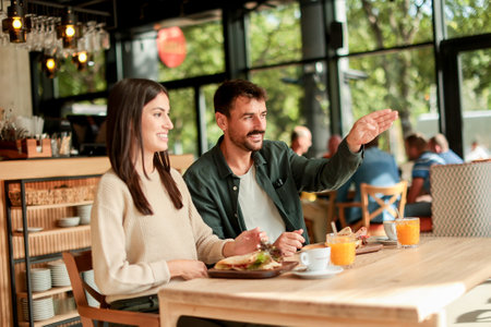 Couple shares laughter and delicious food while sitting at a wooden table in a bright cafe, surrounded by greenery and other patrons enjoying their mealsの写真素材