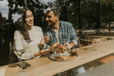 A couple delightfully indulges in slices of pizza at a charming cafÃ©, surrounded by bright, airy...