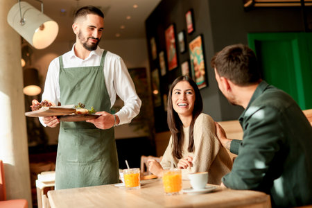 In a lively cafe filled with colorful artwork, a waiter presents fresh vegetarian meals to a smiling couple enjoying their afternoon together over refreshing drinksの写真素材
