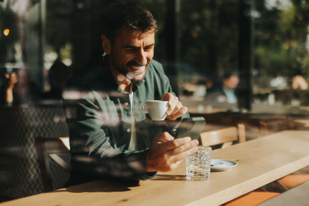 Sitting at a wooden table, a man enjoys his coffee while using mobile phone and gazing out a window, capturing the essence of solitude and reflection in a warm cafe atmosphereの写真素材