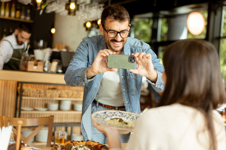 Joyful couple sits at a charming cafe table, smiling as one captures the moment on a smartphone,...