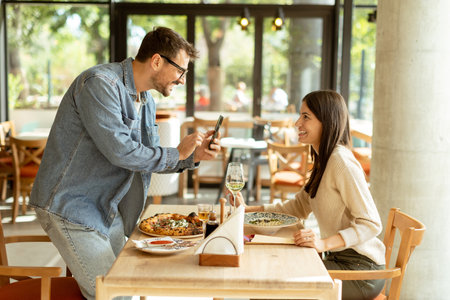 Joyful couple sits at a charming cafe table, smiling as one captures the moment on a smartphone, surrounded by delicious food and a sunny ambianceの写真素材