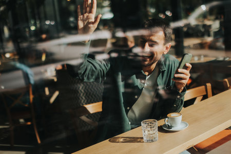 Sitting at a wooden table, a man enjoys his coffee while using mobile phone and gazing out a window,...