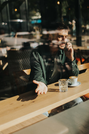 Sitting at a wooden table, a man enjoys his coffee while using mobile phone and gazing out a window, capturing the essence of solitude and reflection in a warm cafe atmosphereの写真素材