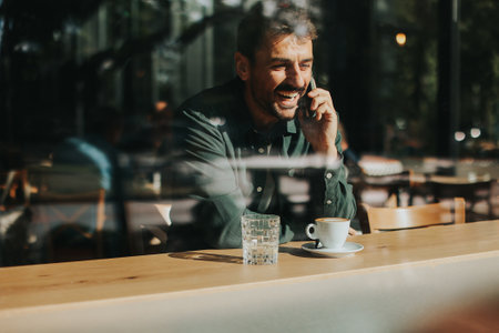 Sitting at a wooden table, a man enjoys his coffee while using mobile phone and gazing out a window, capturing the essence of solitude and reflection in a warm cafe atmosphereの写真素材