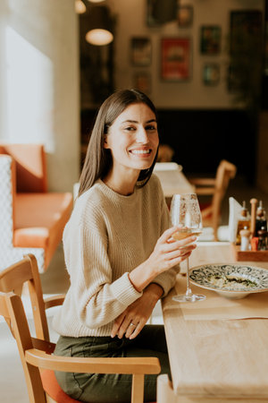 A young woman smiles brightly as she raises a glass of white wine in celebration, savoring her delicious meal in a cozy, modern restaurant filled with vibrant decor.の写真素材
