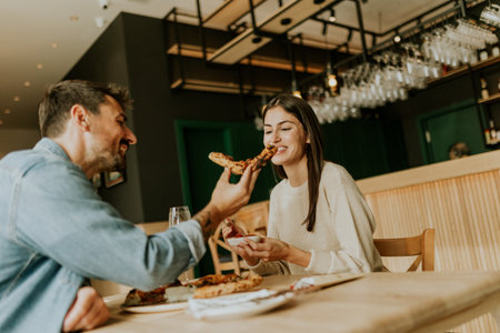 A joyful couple shares a moment at a rustic restaurant, savoring slices of pizza while laughing together and enjoying the vibrant atmosphere around them.の写真素材
